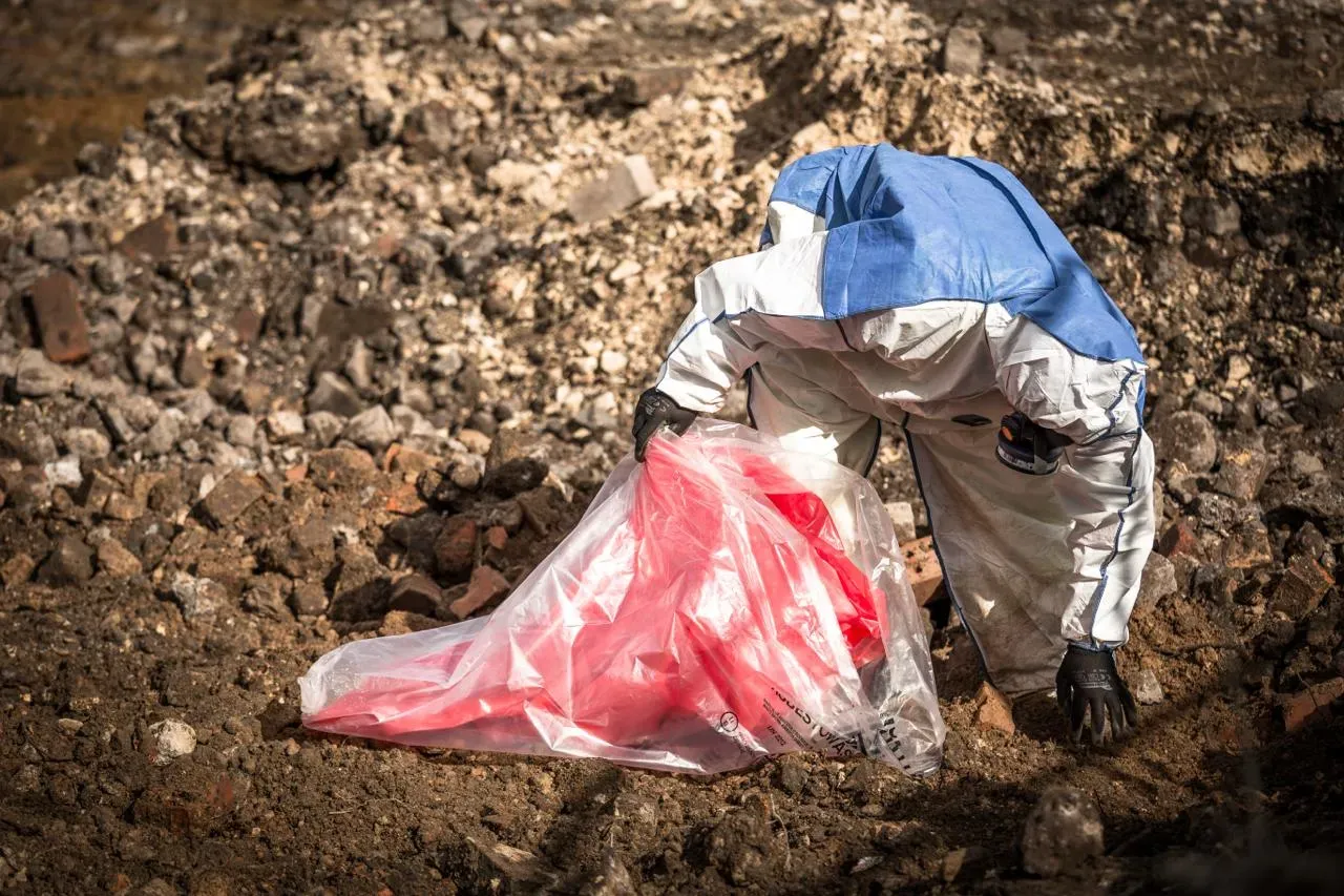 Asbestos removal worker in a protective suit and respirator, carefully bagging hazardous waste material into a clear plastic bag with a red liner on a dusty construction or demolition site.